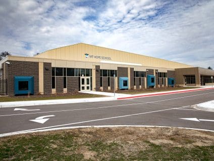 Angled exterior view of Mt. Hope Elementary School showing tan metal panel and dark brick veneer cladding with teal accent window boxes, seen from the parking lot with the “Mt. Hope School” sign visible